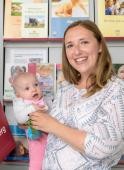 Mutter mit Baby im Familienbüro (Foto: Rothe) Eine Mutter mit Baby auf dem Arm steht vor einem Regal mit Informationsbroschüren im Familienbüro und lacht in die Kamera. (Foto: Rothe)