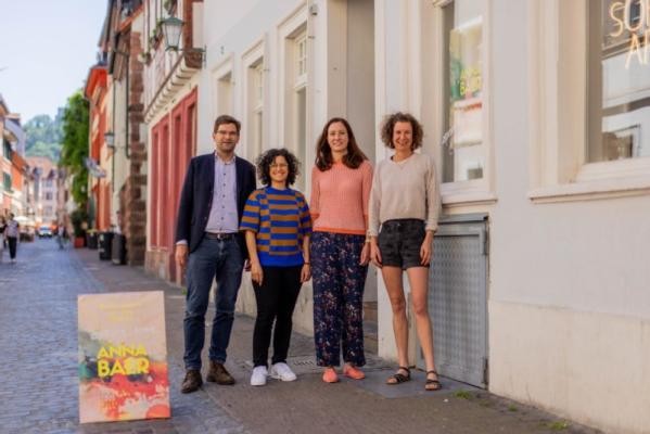 Die Künstlerin Anna Baer (rechts) zeigt in der Unteren Straße 2 ab Sonntag, 19. Mai 2024, ihre Ausstellung „Built Close to the Water“. Drei Personen, die neben einem kleinen Werbeplakat in der Unteren Straße stehen.