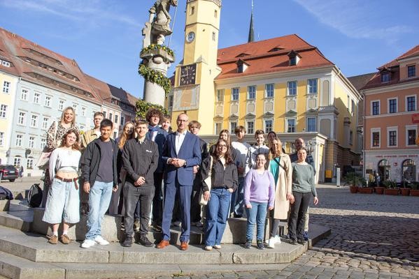 Der Oberbürgermeister von Bautzen, Karsten Vogt (Mitte), traf junge Menschen aus Heidelberg, Bautzen und der umliegenden Region sowie Jelenia Góra zum Austausch – darunter die Heidelberger Jugendgemeinderäte Rafael Sison (vorne, 2. von links), Annika Bischofberger (2. Reihe, 3. von links) und Leni Kischkel (vorne rechts) sowie Leo vom St. Raphael-Gymnasium (2. Reihe, 5. von links). (Foto: Stadt Bautzen) Eine Gruppe junger Menschen mit einem mittelalten Herrn in Anzug stehen vor einem Brunnen in malerischer Altstadtkulisse.