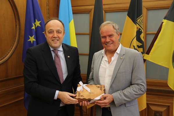 Oberbürgermeister Eckart Würzner (rechts) empfing im Rathaus den ukrainischen Generalkonsul Yurii Nykytiuk. (Foto: Stadt Heidelberg) Zwei Herren in Anzug posieren mit einem Geschenk, hinter ihnen die Flaggen der EU, Ukraine, Deutschlands und Baden-Württembergs.