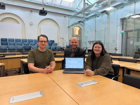 Freuen sich über die erfolgreiche Einführung der Protokollierung mithilfe von Künstlicher Intelligenz (von links): Sascha Verhoeven, Team Innovation im Referat des Oberbürgermeisters, Stefan Lenz, Leiter der Sitzungsdienste, und Michelle Eisel, Sachgebietsleiterin bei den Sitzungsdiensten.(Foto: Stadt Heidelberg) Zu sehen sind 3 Personen die im Großen Sitzungsaal sitzen und einen Laptop präsentieren.