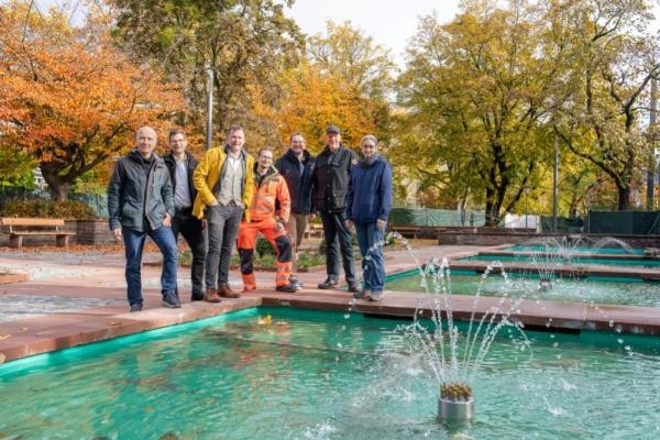 Nach vielen Jahren laufen die Wasserspiele in der Brunnenanlage Kurfürsten-Anlage wieder: Bürgermeister Raoul Schmidt-Lamontain (3. von links) freut sich mit (von links) Volker Schwarz (Landschafts- und Forstamt), Peter Hoffmann (Amt für Stadtentwicklung und Stadtplanung), Florian Scharwatt, Andreas Lippke, Dr. Ernst Baader und Projektleiterin Karen Engelhardt (alle Landschafts- und Forstamt) über die umfassend sanierte Platzfläche. (Foto: Christian Buck) Eine Gruppe von Männern steht am Rand der Brunnenanlage und posiert vor eine laufenden Fontäne.