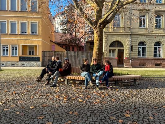 Schon montiert, wurden die Bänke auf dem Römerplatz in Bergheim gut angenommen – eines der letzten Projekte des Programms Mittendrinnenstadt. (Foto: Stadt Heidelberg) Fünf Menschen sitzen auf einer runden Bank die um einen Baum geht. Die Athmosphähre ist herbstlich und die Gebäude im Hintergrund Sind beige.