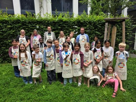 Sind stolz auf das erste internationale Heidelberger Kinderkochbuch (hintere Reihe von links): Rikea Grabs (Bereichsleiterin Ferienbetreuung bei päd-aktiv), Sabine Lachenicht (Leiterin Amt für Umweltschutz, Gewerbeaufsicht und Energie), Jens Katzenberger (Vorstand bei päd-aktiv), Barbara von der Heyden (Digitalisierung/Übergang Kita-Schule beim Amt für Schule und Bildung) und Thorsten Erbismann (Agenda-Büro beim Amt für Umweltschutz, Gewerbeaufsicht und Energie). (Foto: Stadt Heidelberg/päd-aktiv) Kinder in Schürzen.