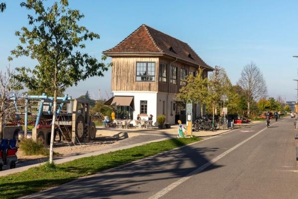 Das Stellwerk an der Bahnstadt-Promenade gilt als wichtiges technisches Kulturdenkmal der regionalen Verkehrsgeschichte. 1914 erbaut, erinnert es an die bewegte Vergangenheit der Bahnstadt, als auf dem Areal des früheren Güter- und Rangierbahnhofs noch mit Wasserdampf betriebene Lokomotiven fuhren. (Foto: Christian Buck) Zweistöckiges Haus mit Terrasse, daneben Spielplatz und Fahrräder an einer sonnigen Straße mit Radfahrern.