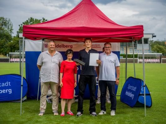 Heidelbergs Jugendsportler des Jahres 2025: Der Schwimmer Morné van Rooyen vom SV Nikar Heidelberg mit (von links) Dr. Peter Schlör, Vorsitzender des Sportkreises Heidelberg, Bürgermeisterin Stefanie Jansen und Gert Bartmann, Leiter des Amtes für Sport und Gesundheitsförderung. (Foto: Tobias Dittmer) Erstplatzierter im Schwimmen Morné van Rooyen wird vor einem Zelt geehrt.