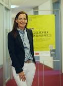 Ines Hotz, Bereichsleiterin Firmen- und Gewerbekunden bei der Sparkasse Heidelberg. (Foto: Sparkasse Heidelberg) Bild von Ines Hotz. Sie steht vor einem Plakat, das für den Gründungspreis wirbt.