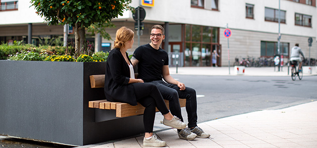 Zwei Menschen unterhalten sich in der Poststraße auf einer Bank.