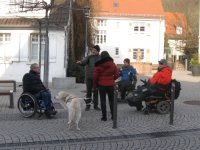 Mehrere Personen und ein Blindenhund stehen auf einem Platz, darunter drei Rollstuhlfahrer